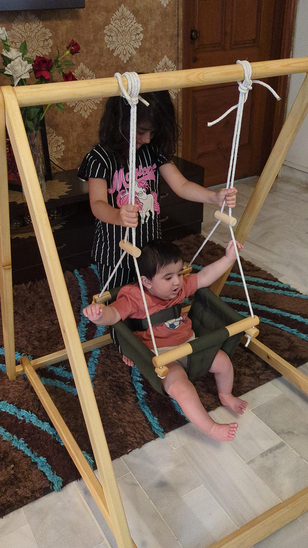 Two children playing on a wooden swing set indoors.
