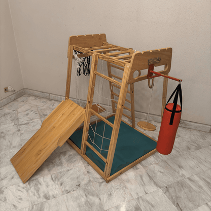 A wooden jungle gym in a room featuring slide, boxing bag, swing, monkey bar, rope ladder