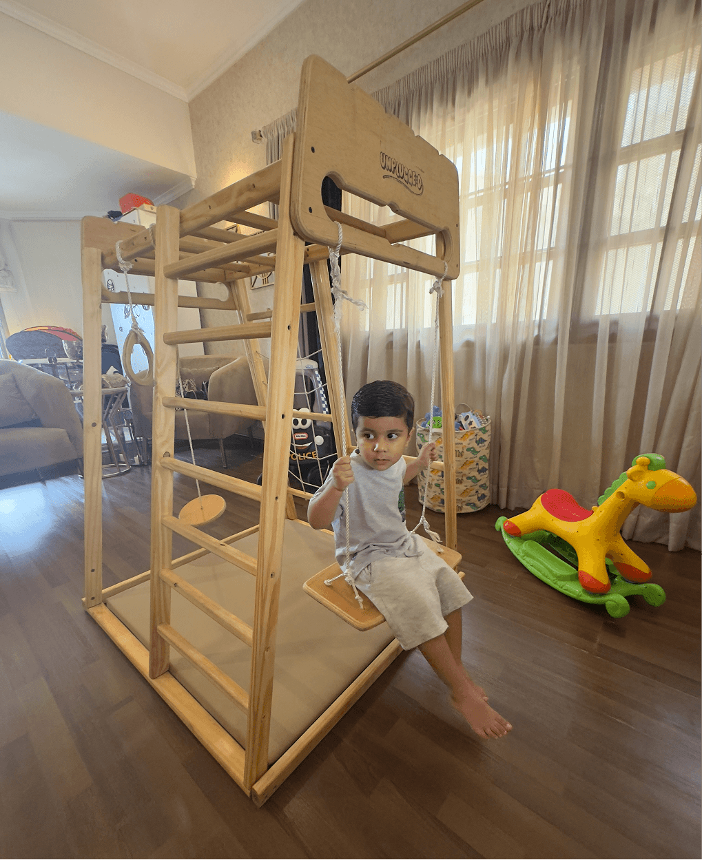 Child swinging on a wooden jungle gym in a room with toys around
