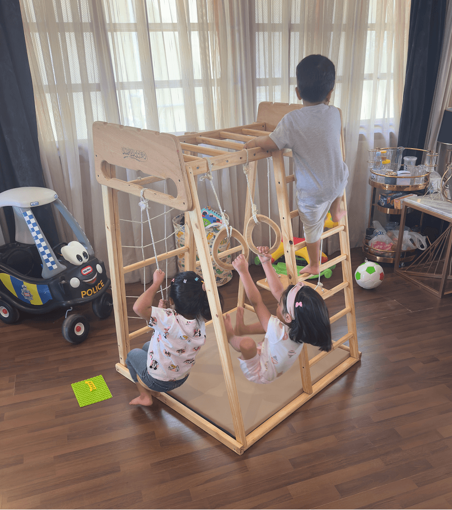 Children playing on a jungle gym, climbing on a ladder and swinging and hanging from gymnastic rings in a room with toys around.