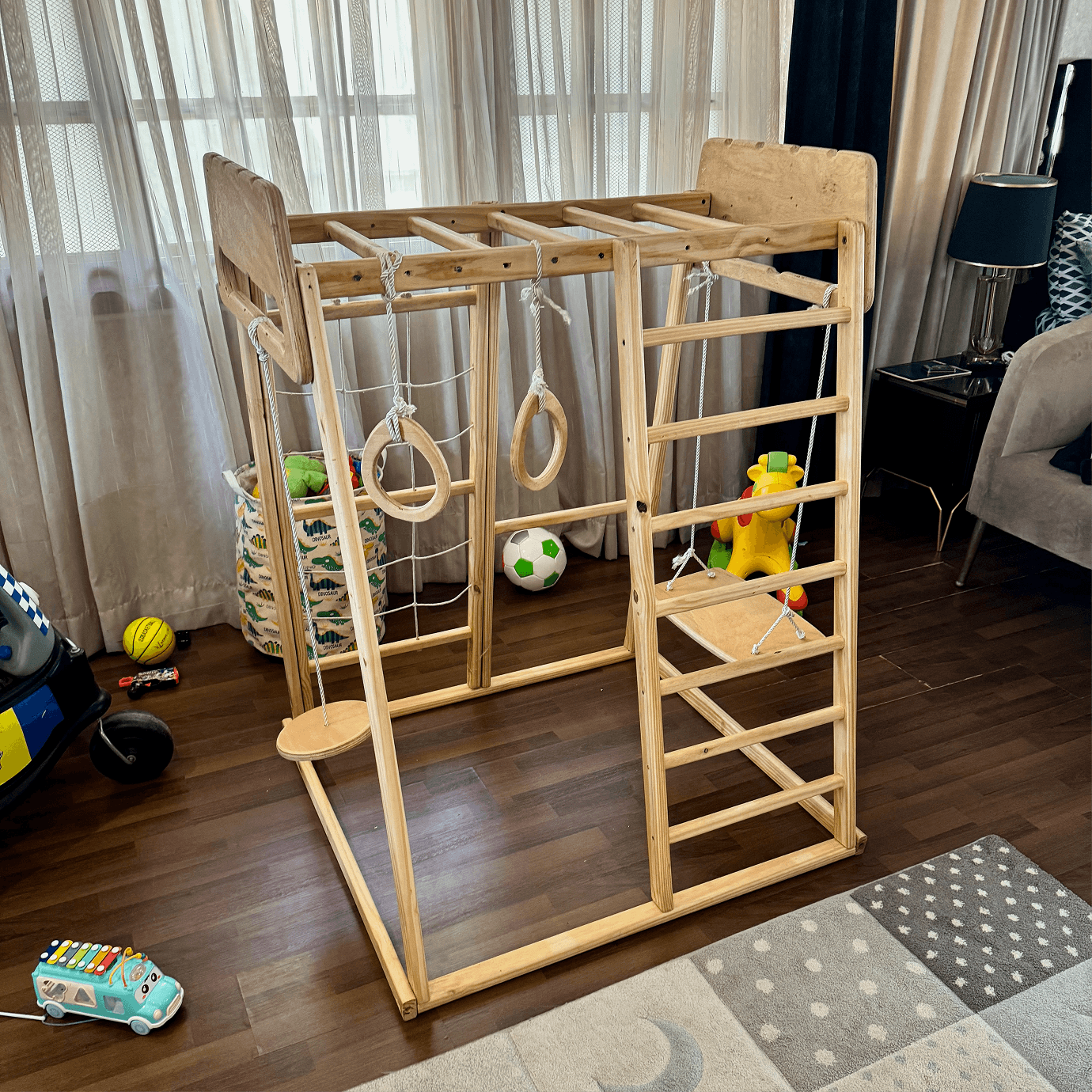 Wooden jungle gym with swing, gymnastic rings, ladder and monkey bar in a room with toys and furniture.