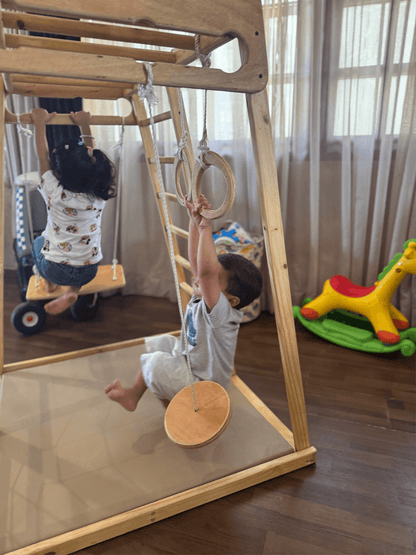 Children playing on a wooden jungle gym with rings, swing and a ladder in a home setting.